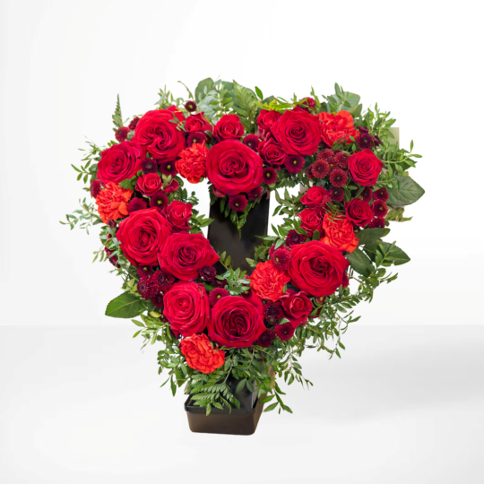 Hjärtformat begravningsarrangemang med röda rosor, mörka blommor och grönt bladverk..