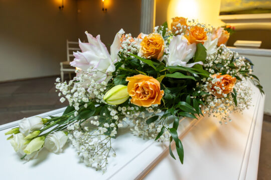 Bouquet of orange roses, white lilies and baby's breath arranged on a white coffin in a softly lit room.